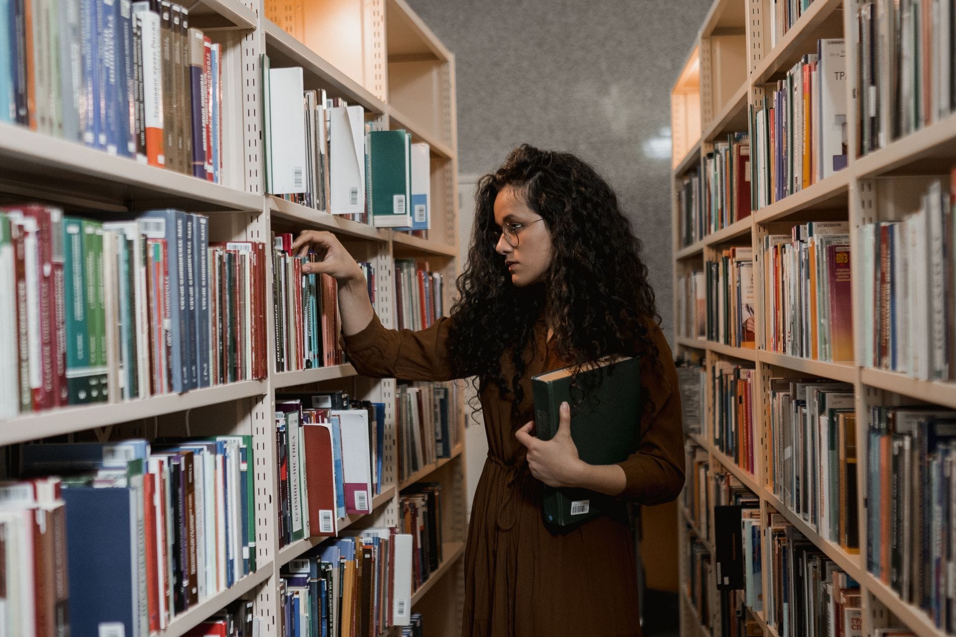 Kobieta w okularach wybiera książkę między regałami w bibliotece