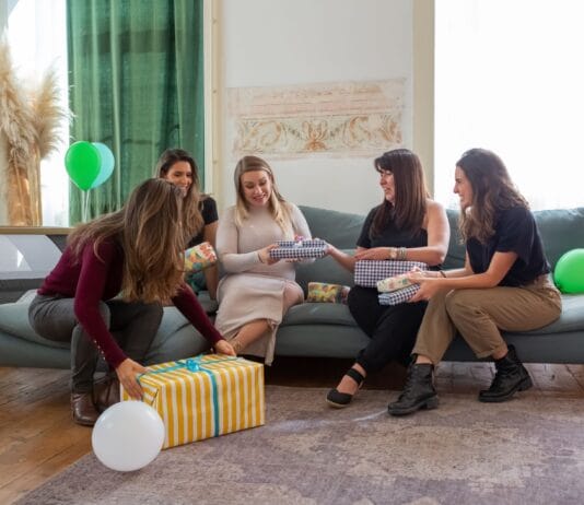 Prezent na baby shower od szefowej i zespołu z pracy – 8 eleganckich propozycji firmowych Kobiety świętujące baby shower i wręczające prezenty w domu